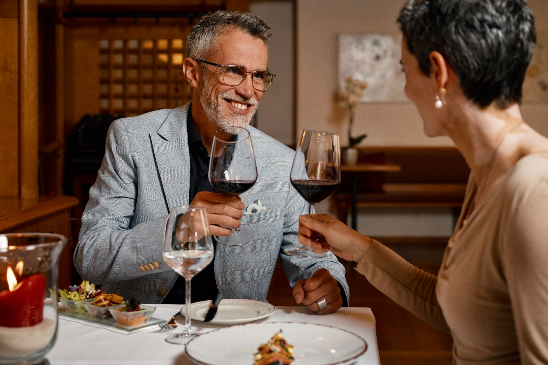 Ein Mann und eine Frau stossen mit Rotweingläsern an. Die Frau sitzt mit dem Rücken zur Kamera, der Mann lächelt sie an. Auf dem gedeckten Tisch stehen Essen und eine brennende Kerze.